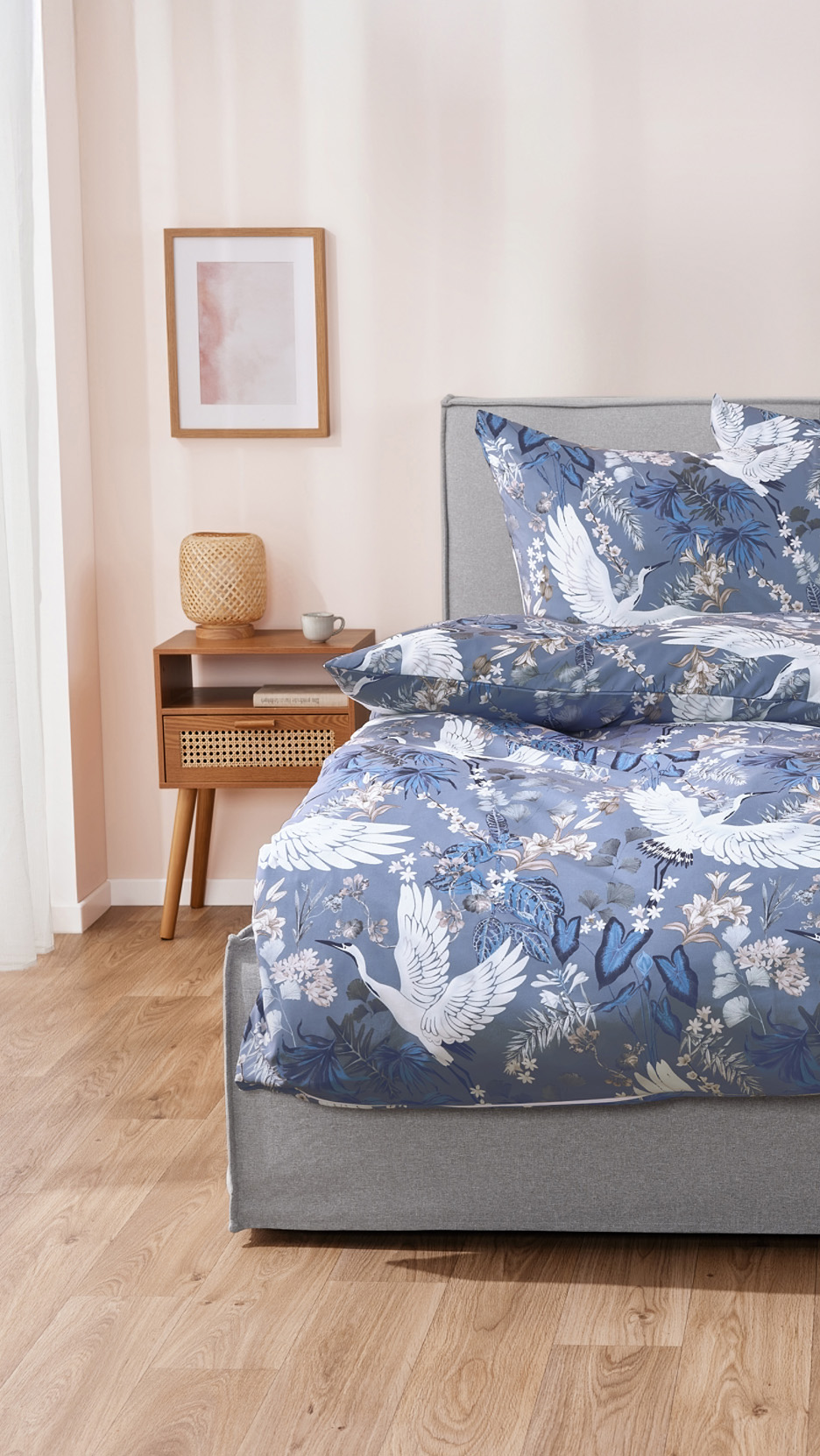 Lenjerie de pat albastră cu model de cocori și plante, pe un pat gri, lângă o noptieră din lemn.