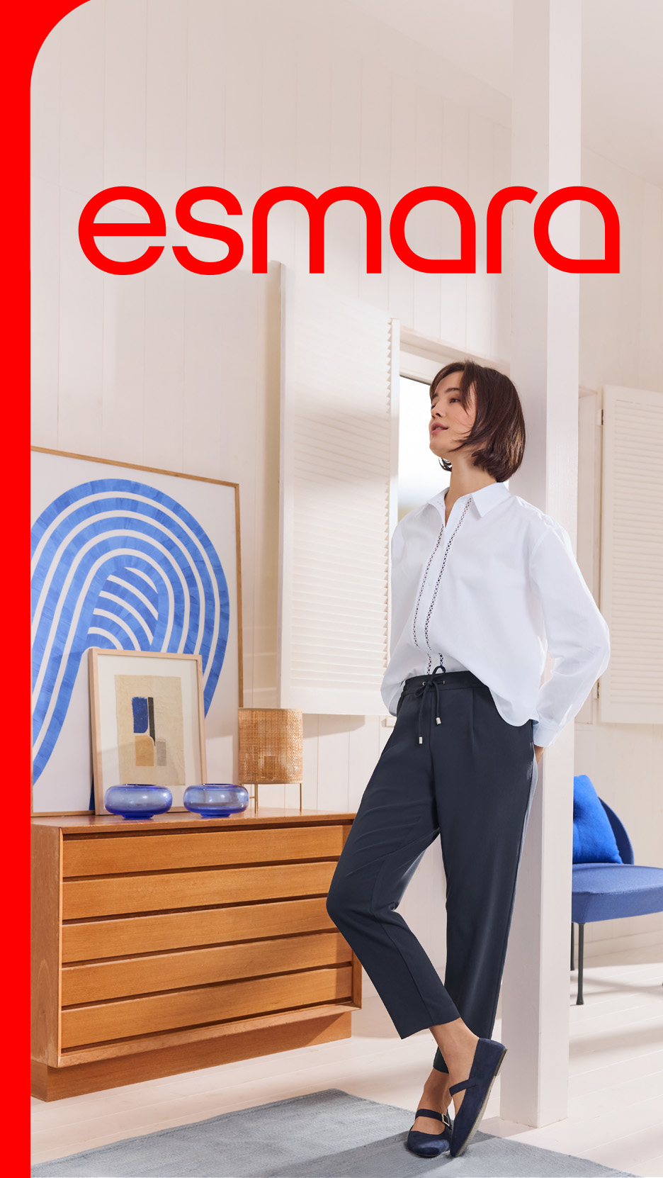 Femeie în cămașă albă și pantaloni bleumarin, sprijinită de perete, cu logo-ul „esmara”.