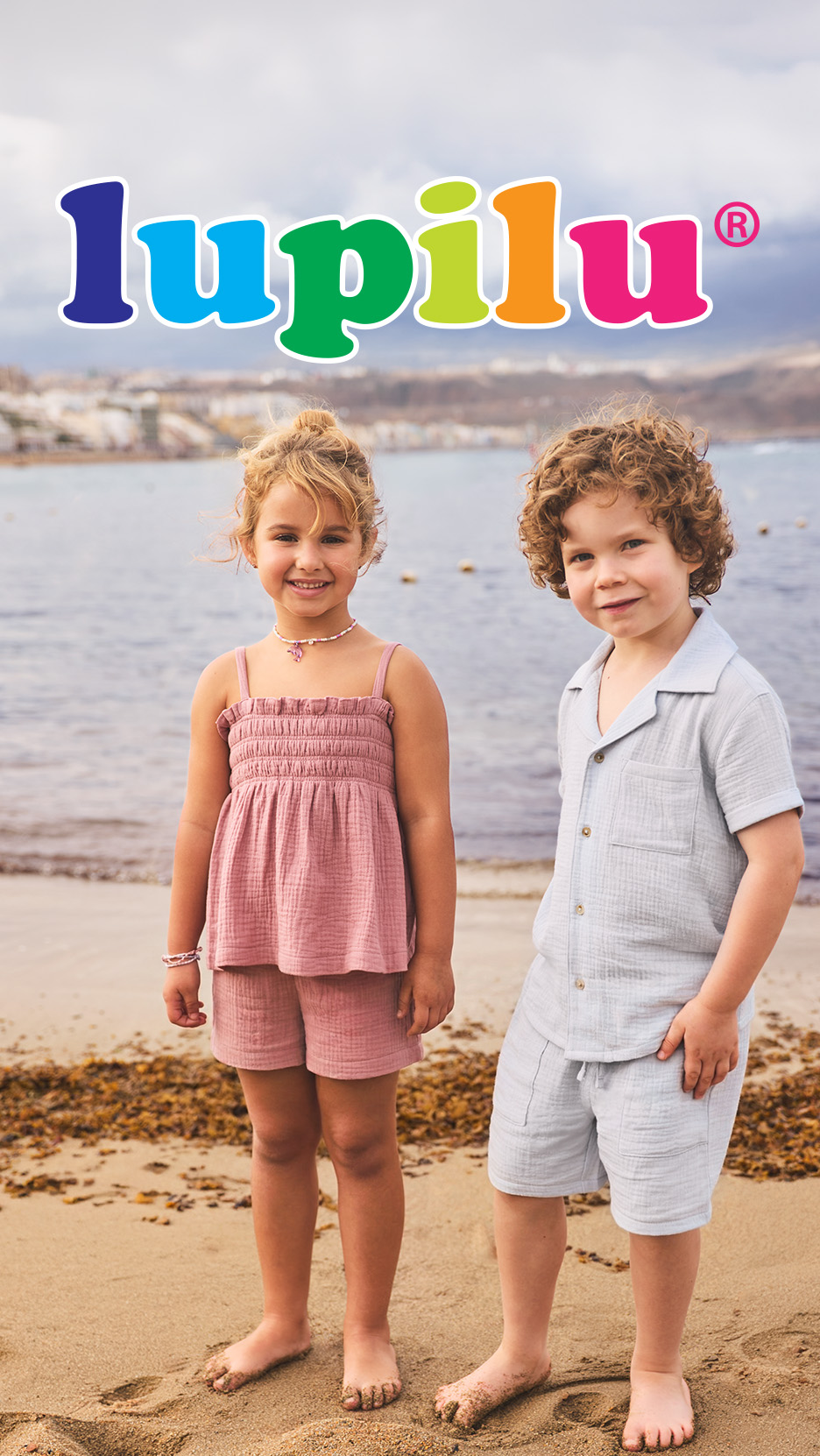 Doi copii zâmbitori pe plajă, purtând haine de vară din bumbac, cu logo-ul Lupilu deasupra.