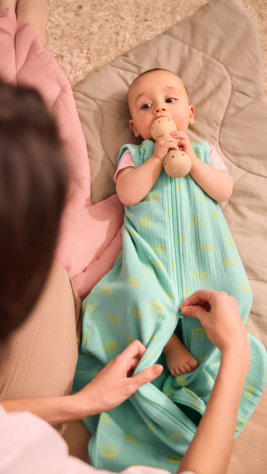 Bebeluș într-un sac de dormit turcoaz cu soare galben, cu o jucărie de lemn în gură.