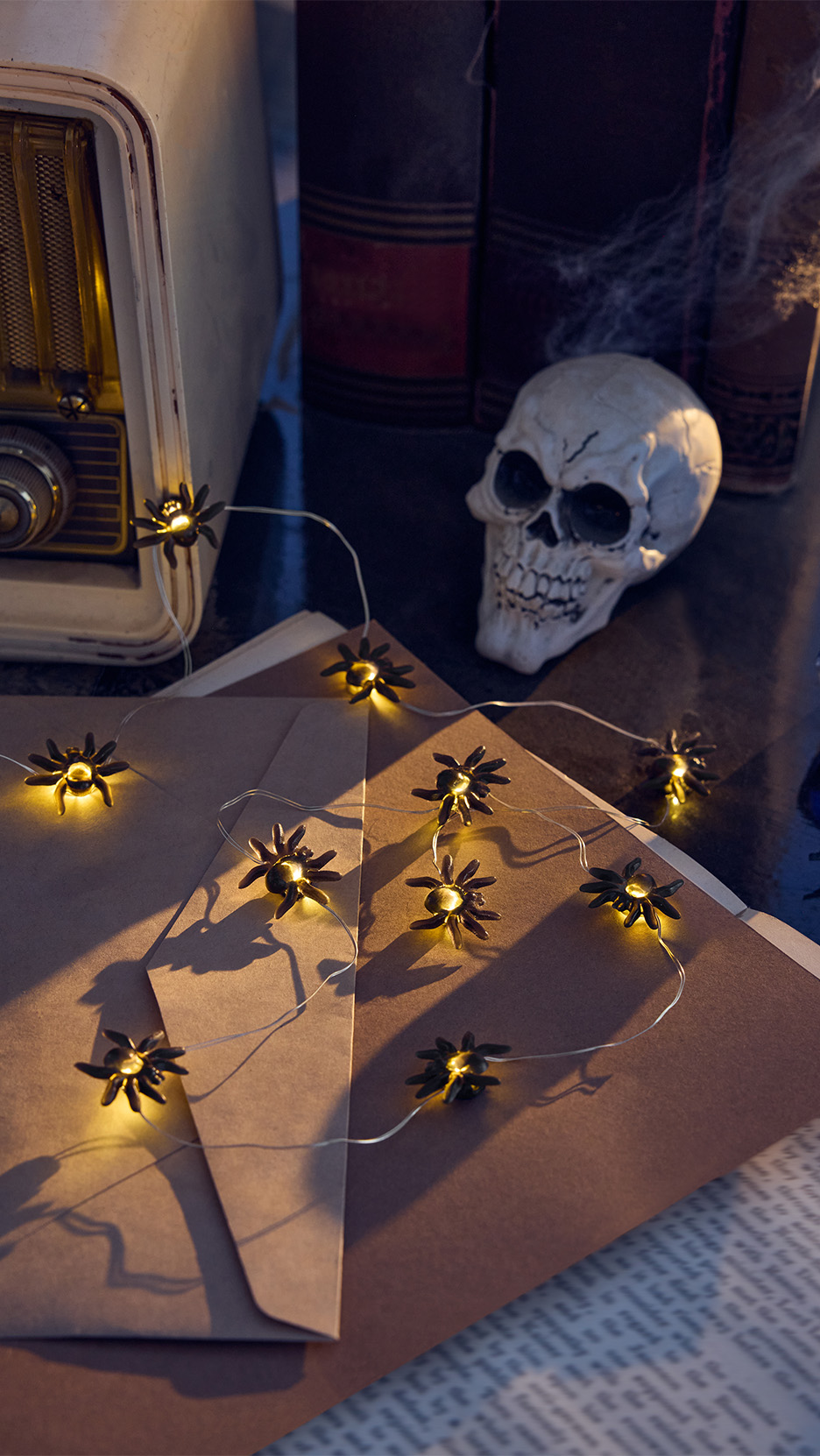 Ghirlandă luminoasă cu păianjeni pe plicuri maro, lângă un craniu decorativ de Halloween