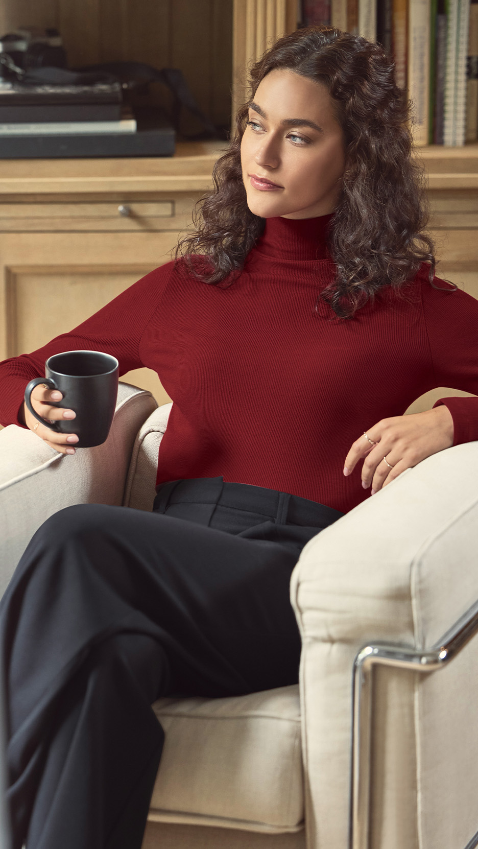 Femeie într-un pulover roșu cu guler înalt și pantaloni negri, ținând o cană neagră.