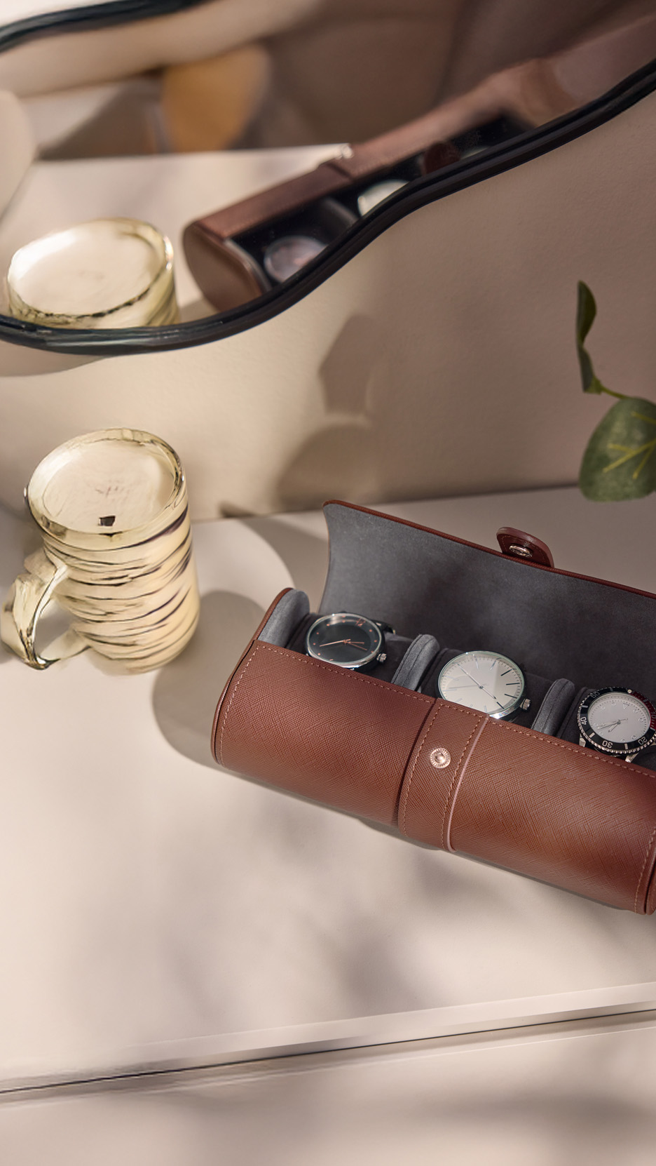 Suport maro din piele pentru ceasuri, cu trei ceasuri, lângă o cană aurie și o oglindă.