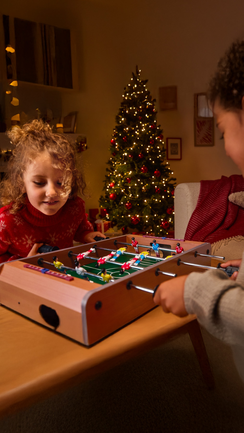 Doi copii se joacă fotbal de masă lângă un brad de Crăciun iluminat.