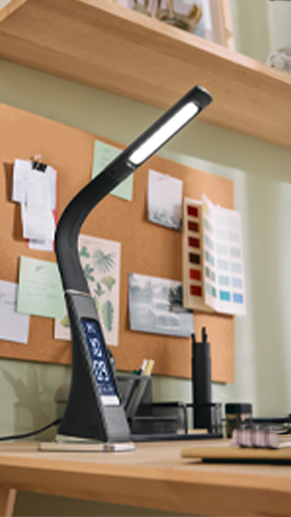 Lampă de birou LED neagră cu afișaj digital pe un birou din lemn, cu panou de plută în fundal.