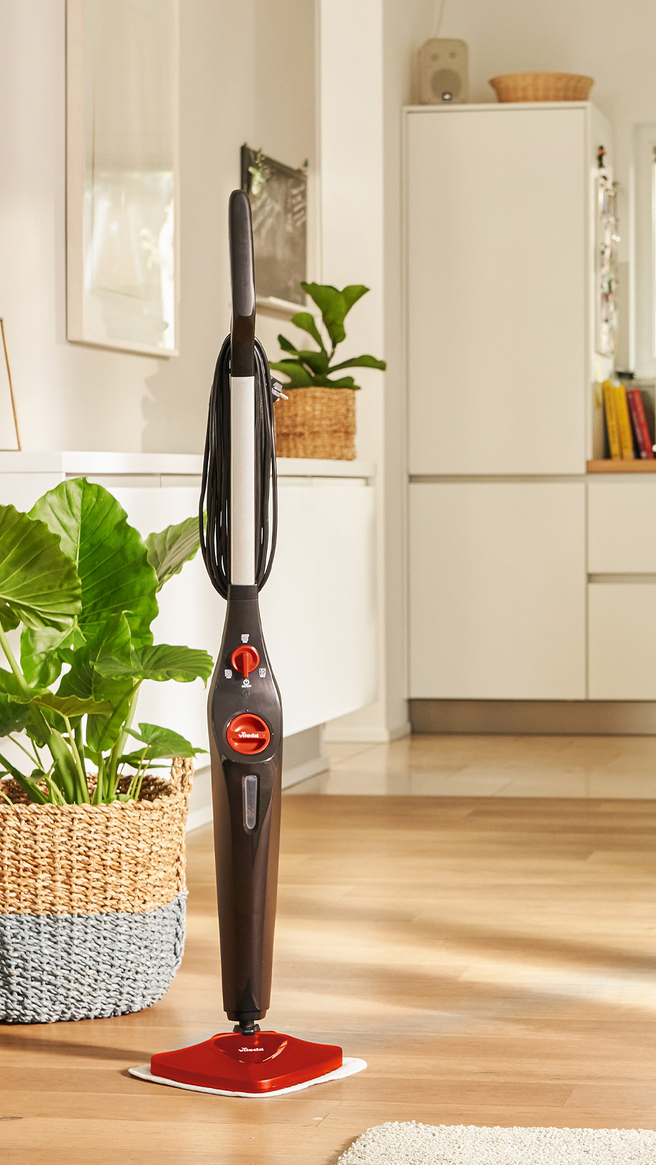 Mop cu aburi Vileda, roșu și negru, pe o podea din lemn, lângă o plantă mare.