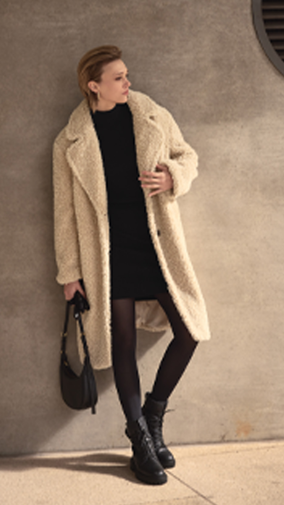 Femeie într-o haină lungă bej, rochie neagră, colanți și cizme, cu o geantă neagră.