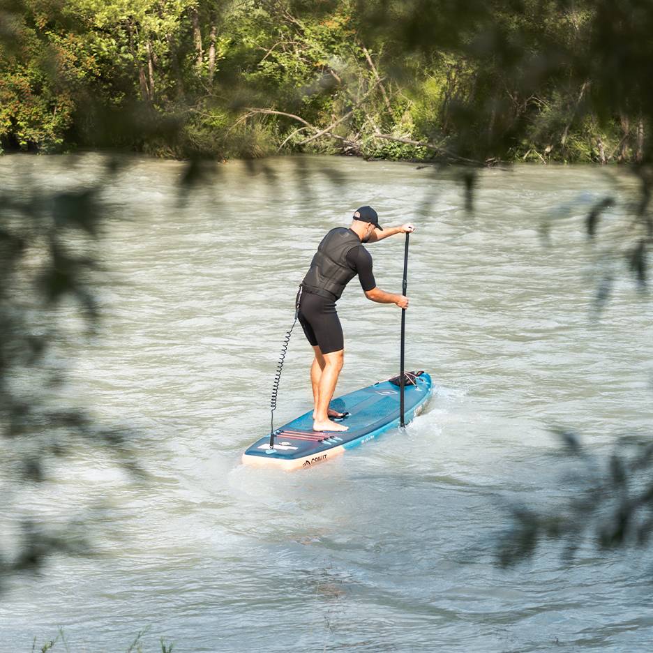 Bărbat în costum de neopren și vestă de salvare vâslind pe o placă de paddleboard albastră pe un râu.