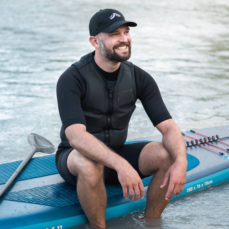 Bărbat zâmbitor cu vestă de salvare și șapcă, stând pe o placă de paddleboard în apă.