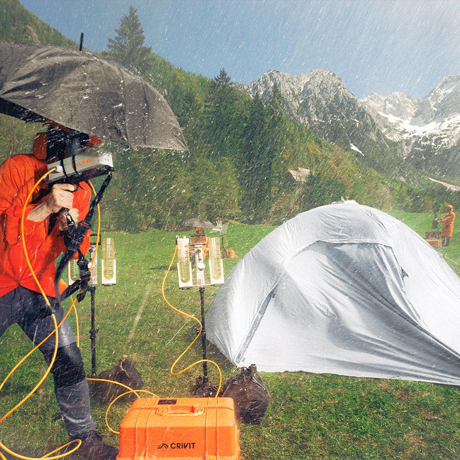 Persoană în echipament de ploaie testând echipament de camping în ploaie, cu munți în fundal.