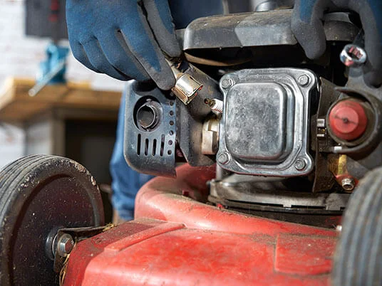 Mâini înmănușate înlocuind bujia unui cositor de gazon roșu.