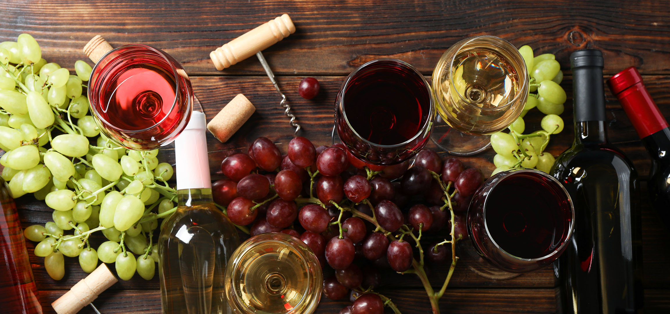 Diverse vinuri, struguri și accesorii de vin pe o masă de lemn.