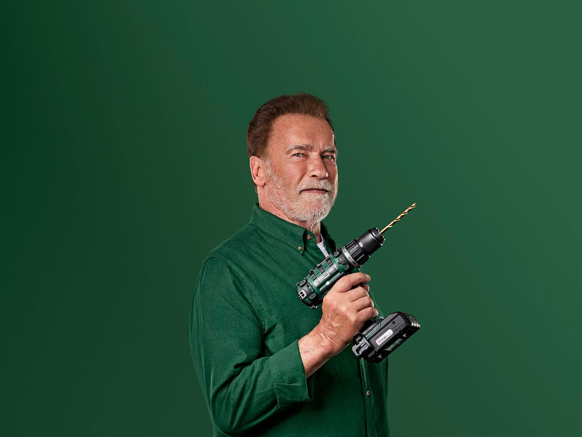 Arnold Schwarzenegger ține o mașină de găurit electrică verde pe un fundal verde.