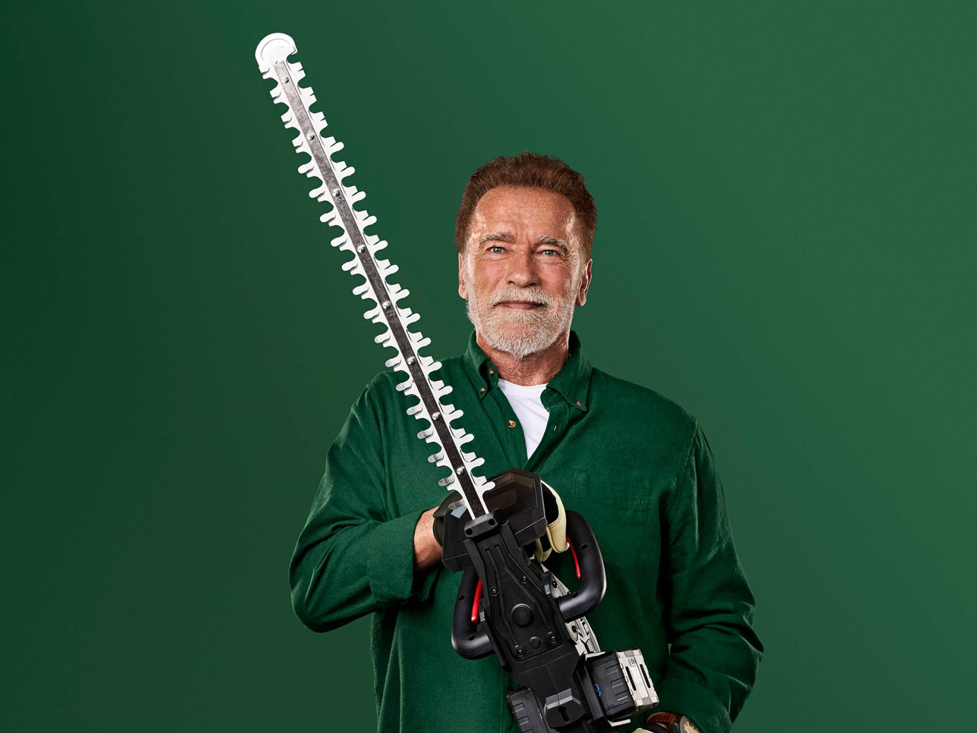 Arnold Schwarzenegger ține un foarfece mare de gard viu pe un fundal verde.