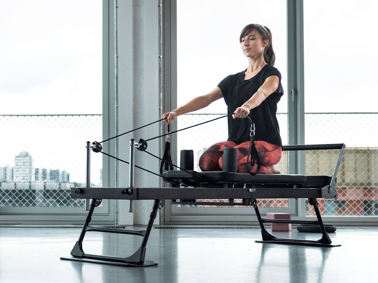 Femeie care face Pilates pe un aparat reformer, cu fundal urban.