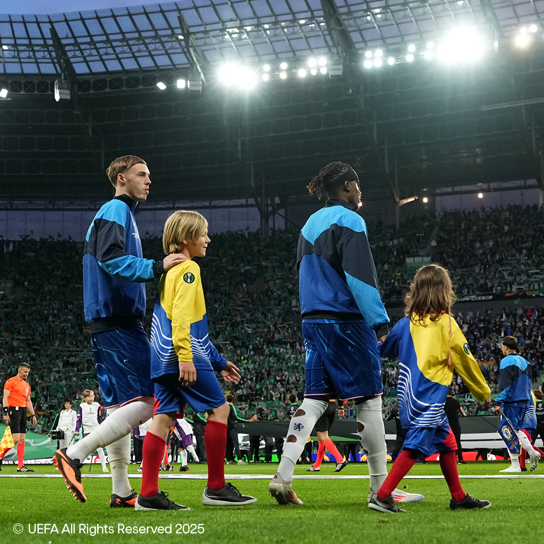 Jucători de fotbal și copii mergând pe terenul unui stadion plin.