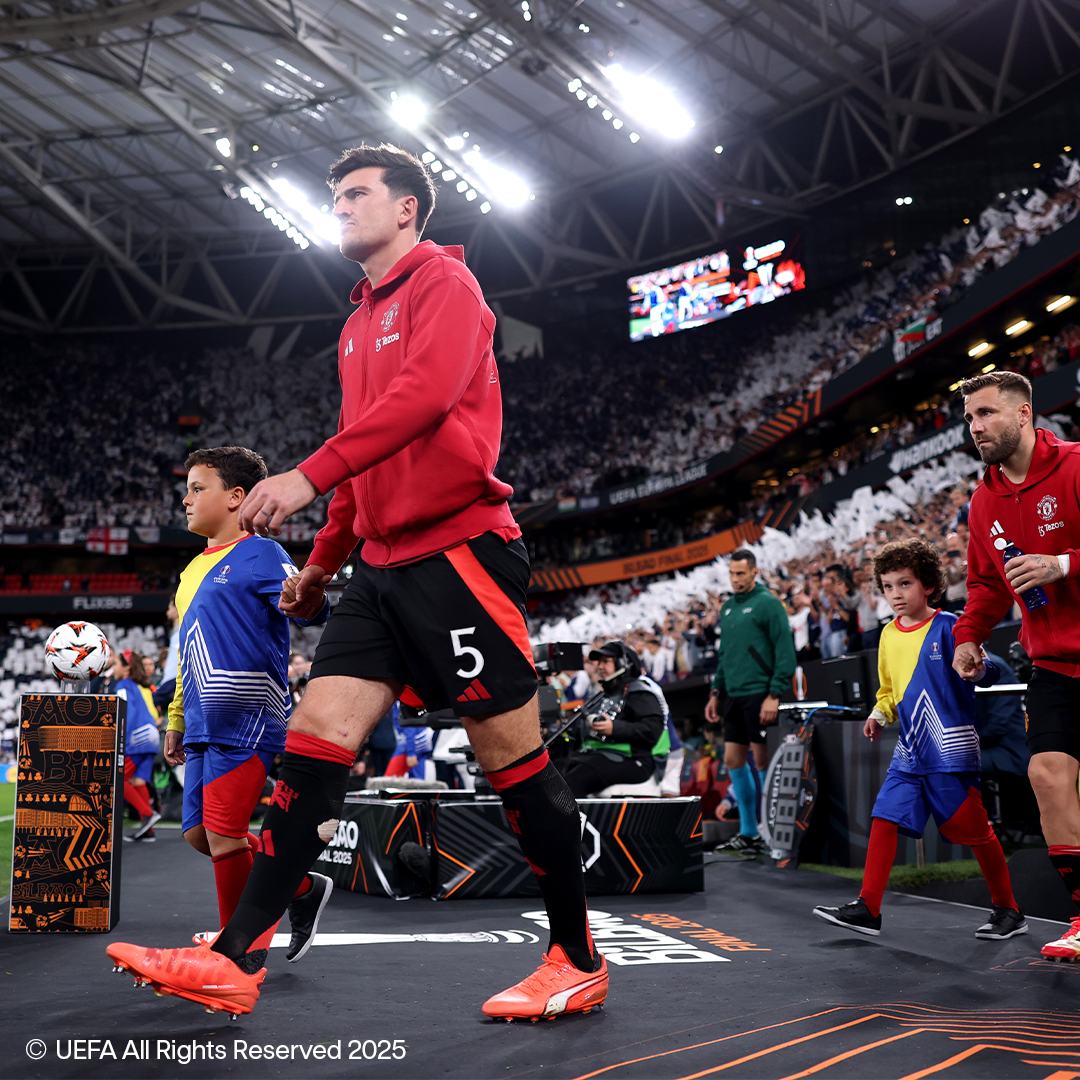 Jucători de fotbal în tricouri roșii și șorturi negre merg pe un teren cu copii, îmbrăcați în haine sport.