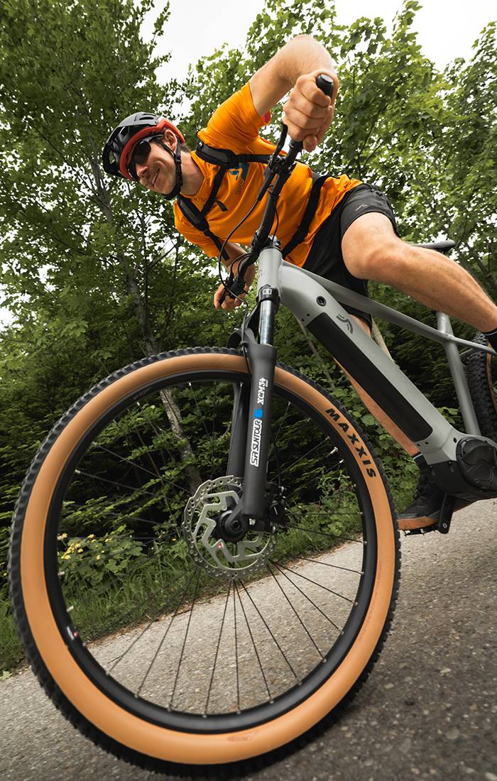 Un bărbat se plimbă cu o bicicletă electrică Maxxis gri prin pădure.