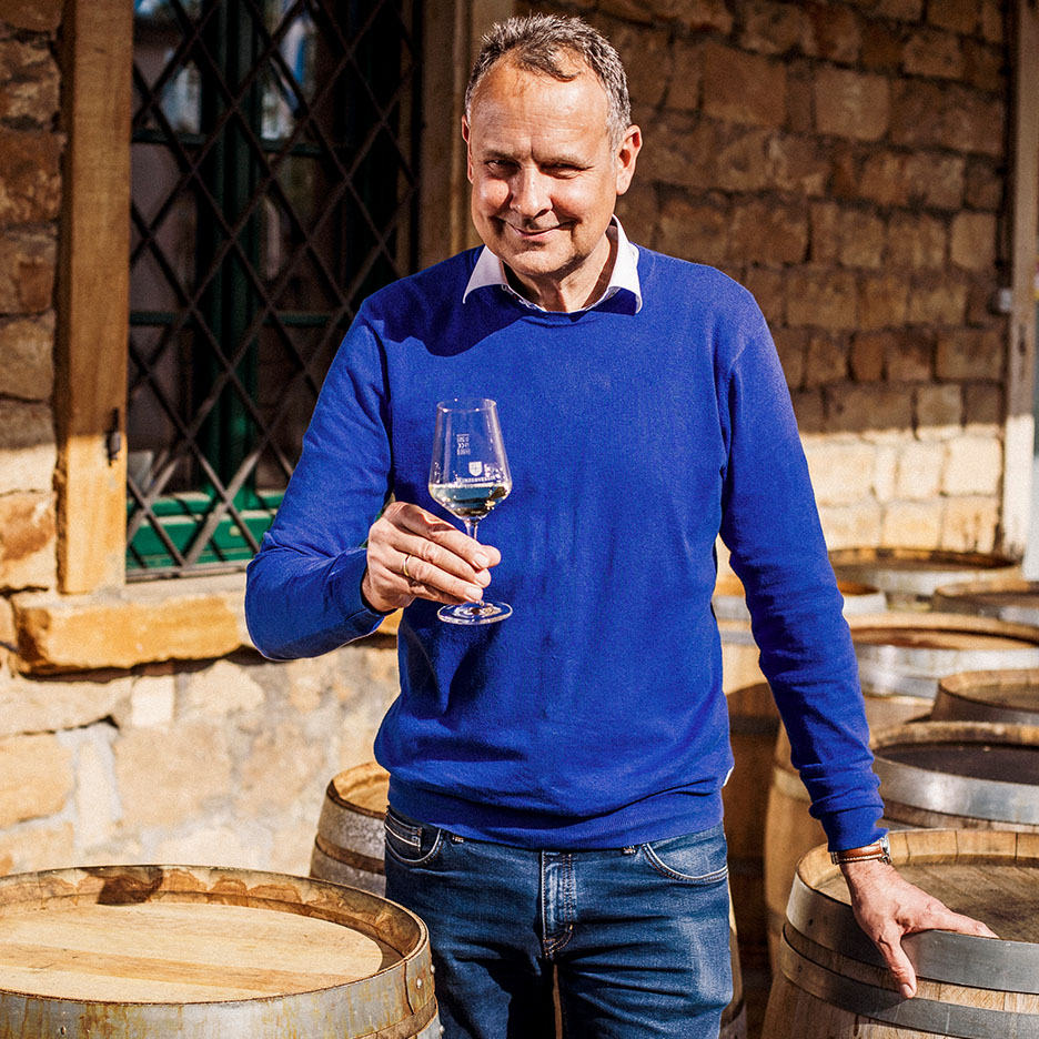 Bărbat zâmbitor în pulover albastru ținând un pahar de vin, cu butoaie de lemn în fundal.