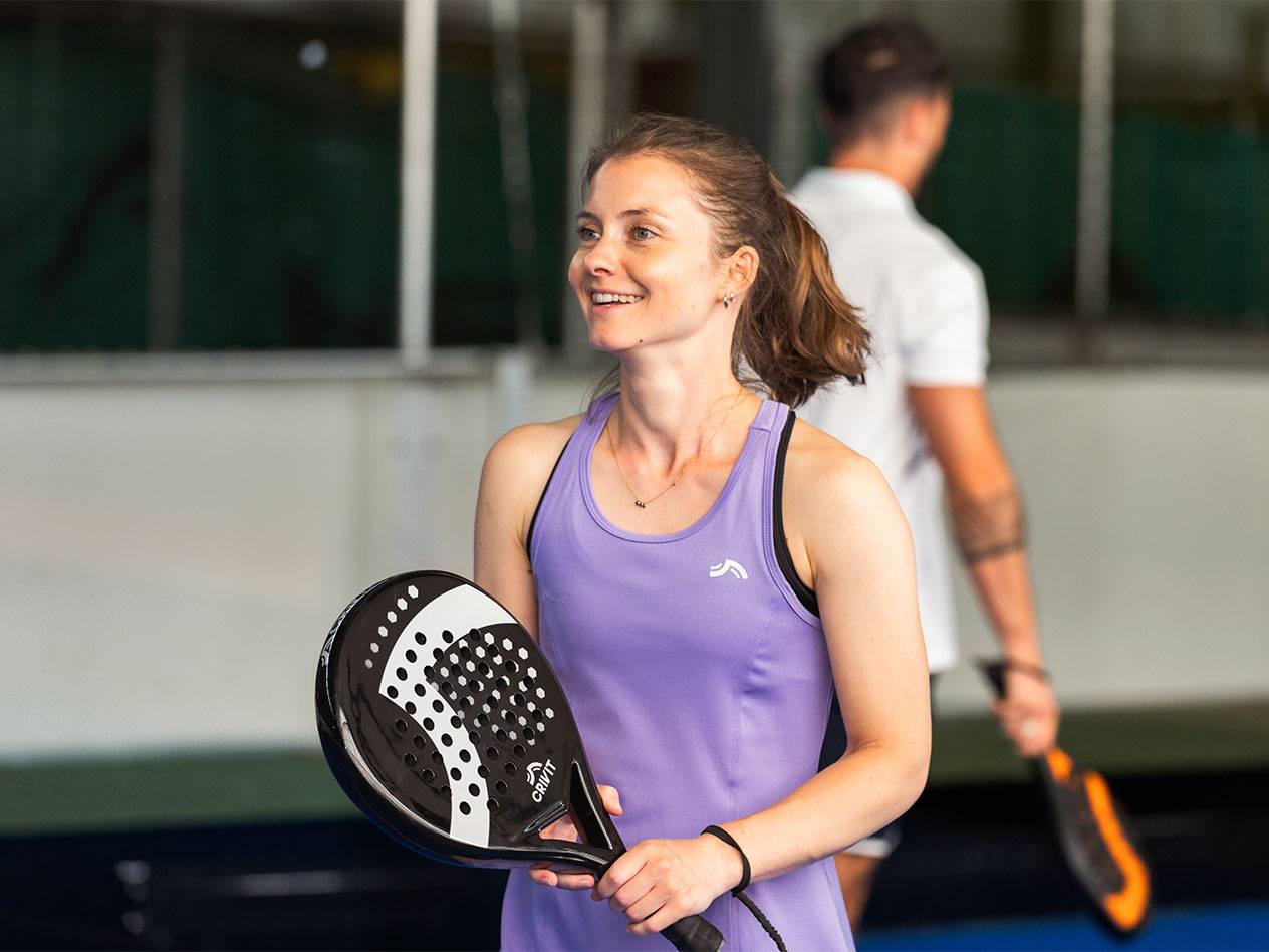 Femeie în rochie sport violetă ținând o rachetă de padel, bărbat în fundal.