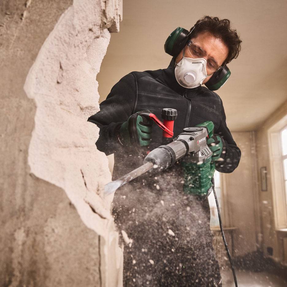 Bărbat cu ciocan demolator și echipament de protecție demolând un perete.