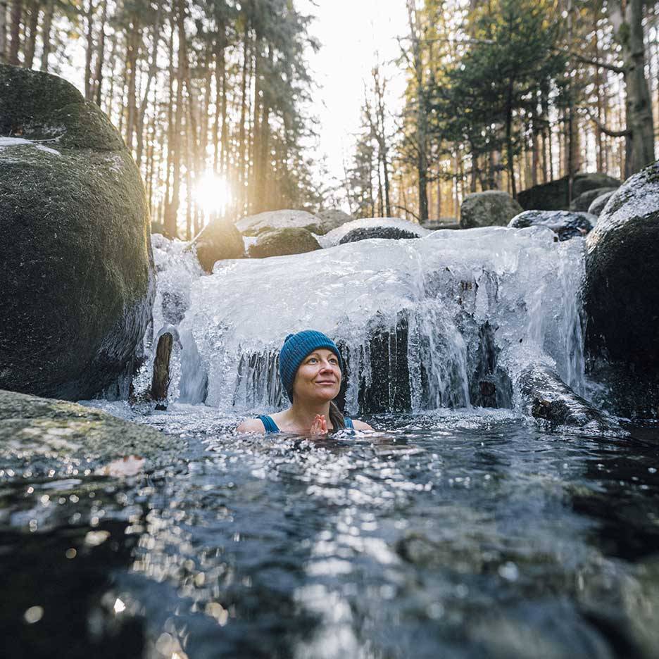 Femeie cu căciulă și costum de baie albastre, bucurându-se de o baie rece într-un râu înghețat.