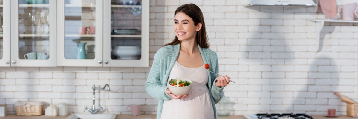 Femeie însărcinată mănâncă o salată în bucătărie.