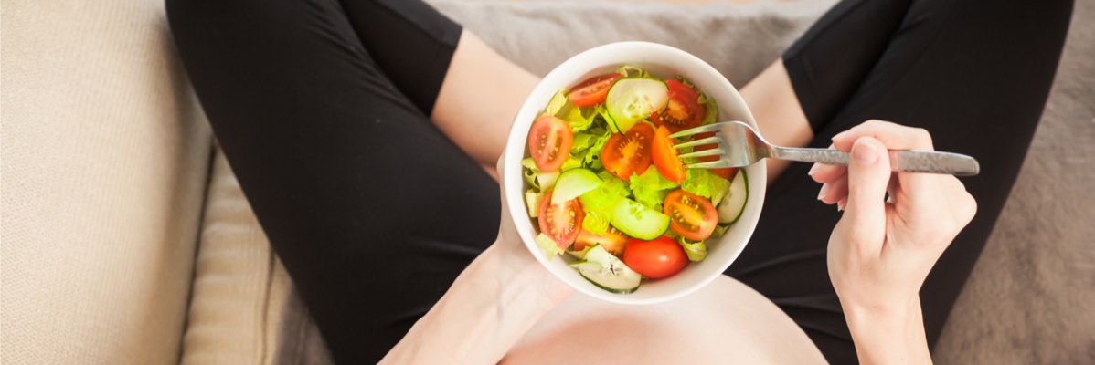 Femeie însărcinată mănâncă o salată cu roșii și castraveți.