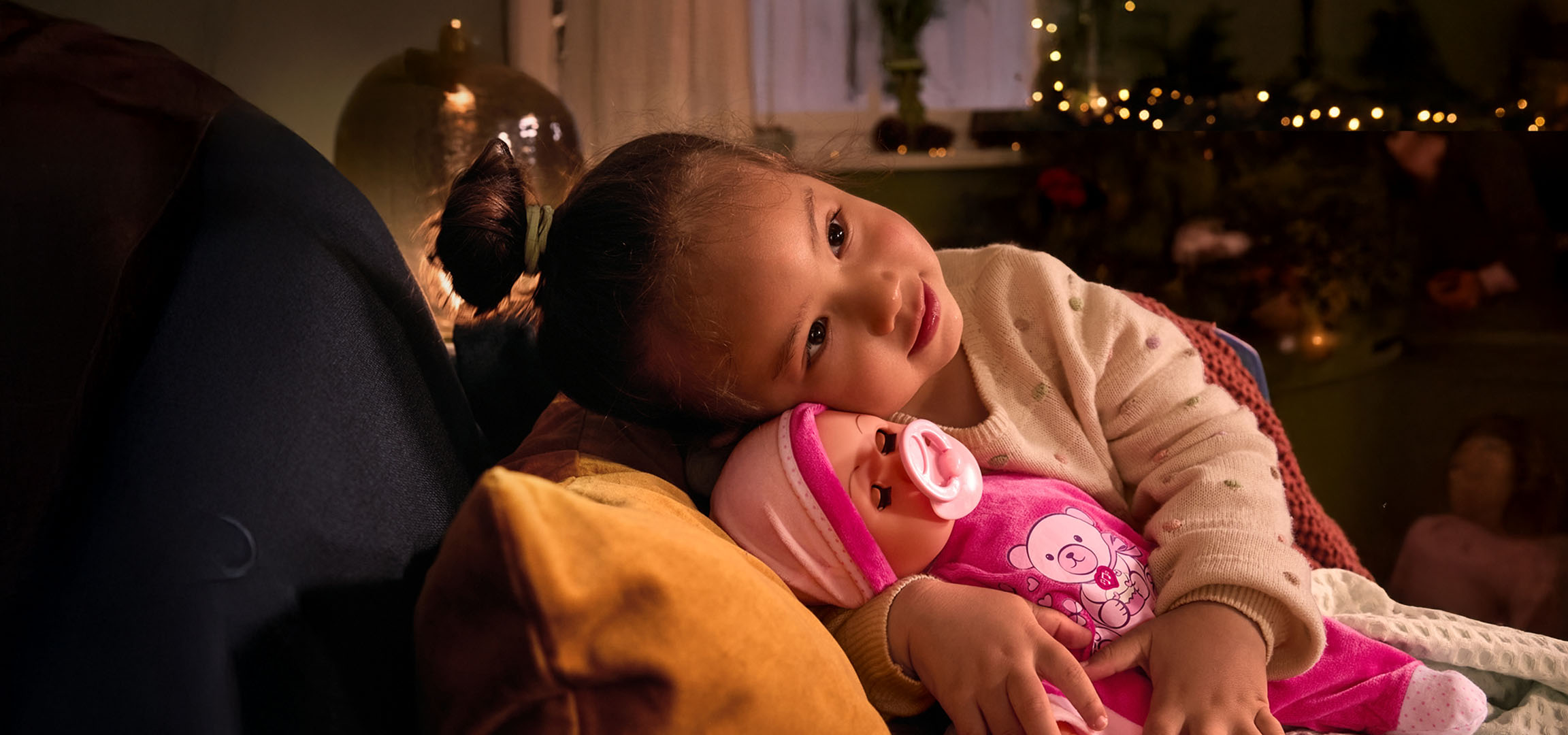 Fetiță cu păpușă și suzetă, așezată confortabil pe o pernă.