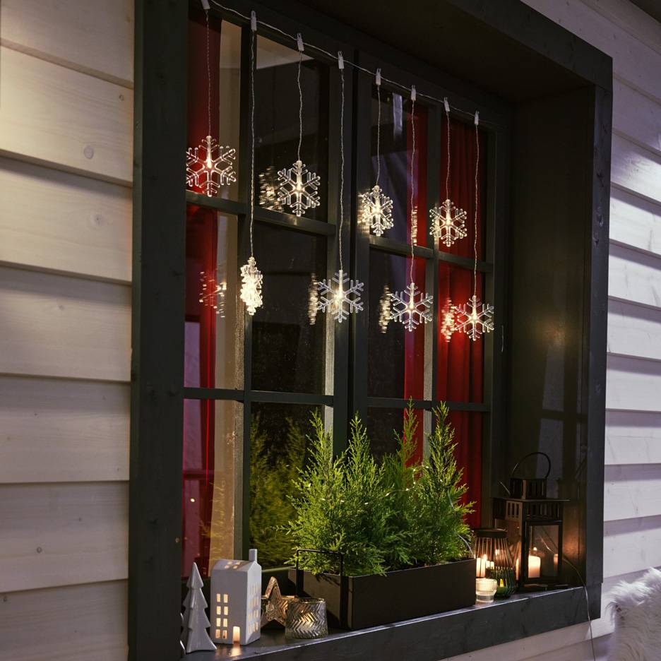 Ghirlandă luminoasă cu fulgi de nea într-o fereastră cu brazi mici și lumânări pe pervaz.