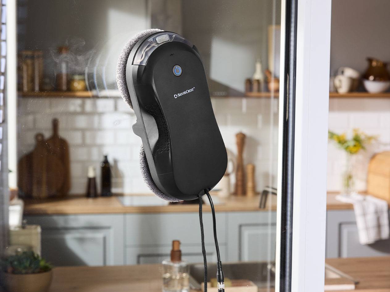 Robot de curățat geamuri Silvercrest pe o fereastră cu o bucătărie în fundal.