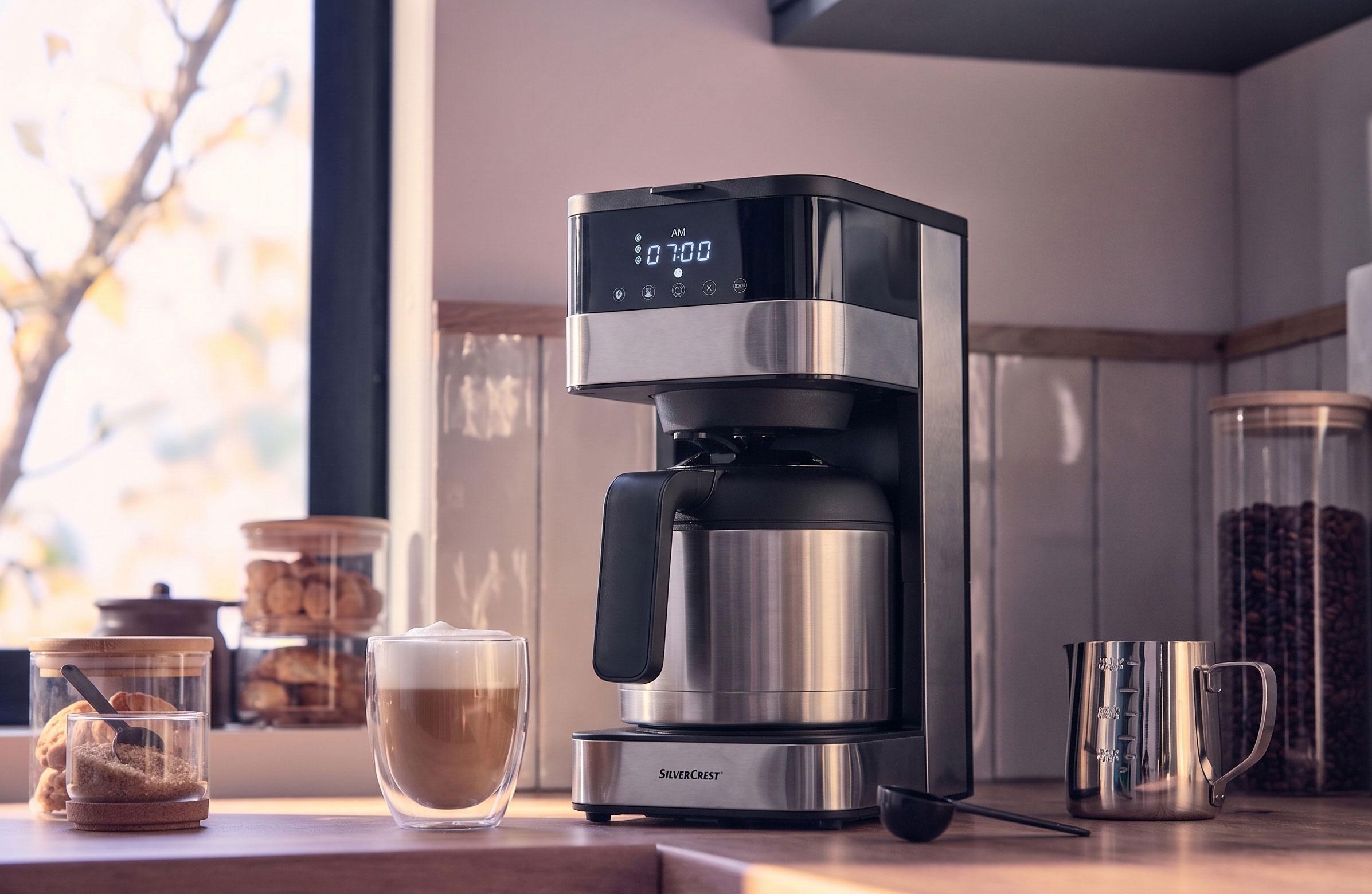 Cafetieră cu carafă termică și afișaj digital care arată 07:00 AM, cu o ceașcă de cafea latte.