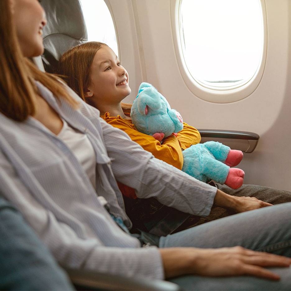 Un copil cu o jucărie de pluș privește pe fereastra unui avion lângă un adult.