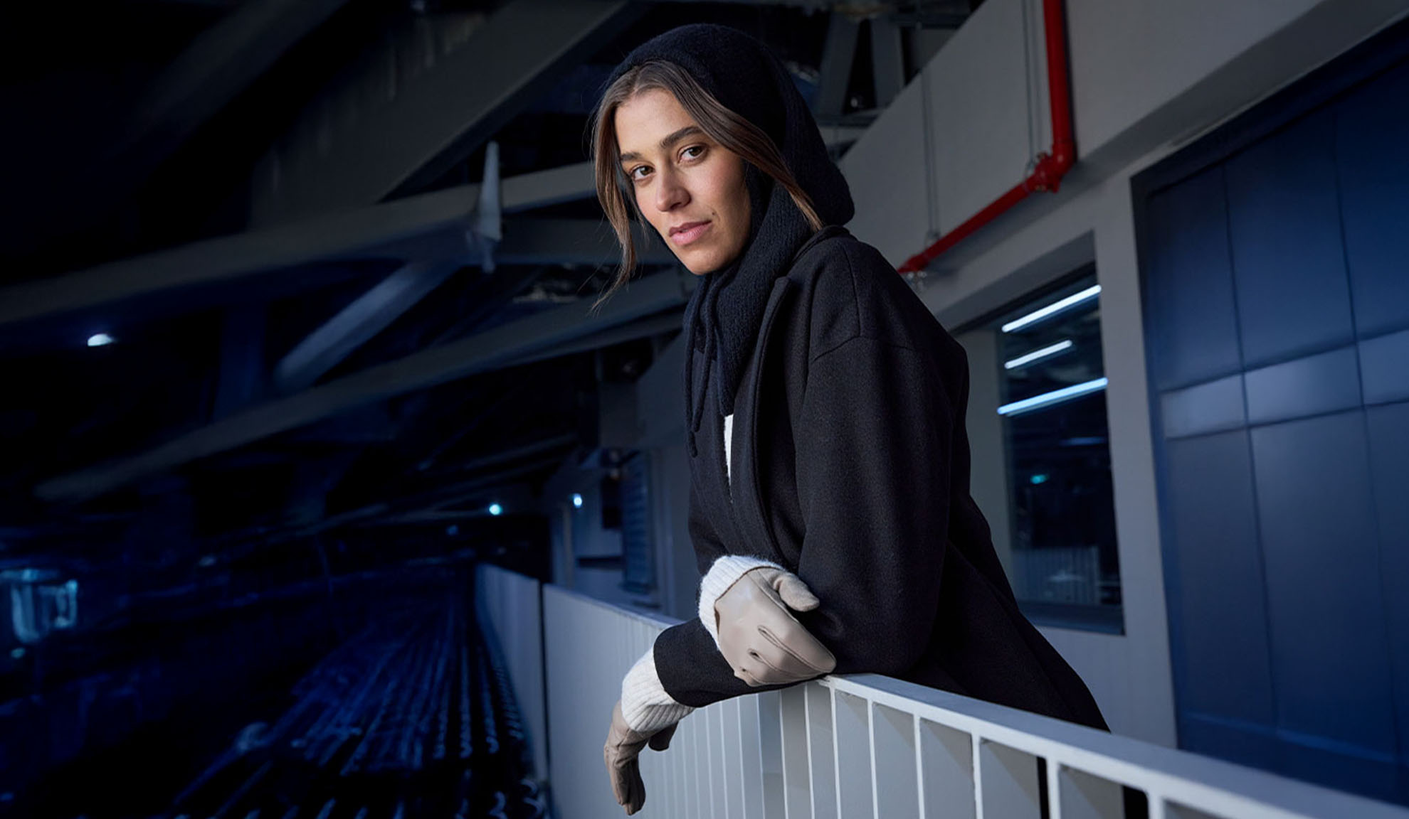 Femeie în palton negru și eșarfă cu glugă, sprijinită de o balustradă într-un cadru modern.