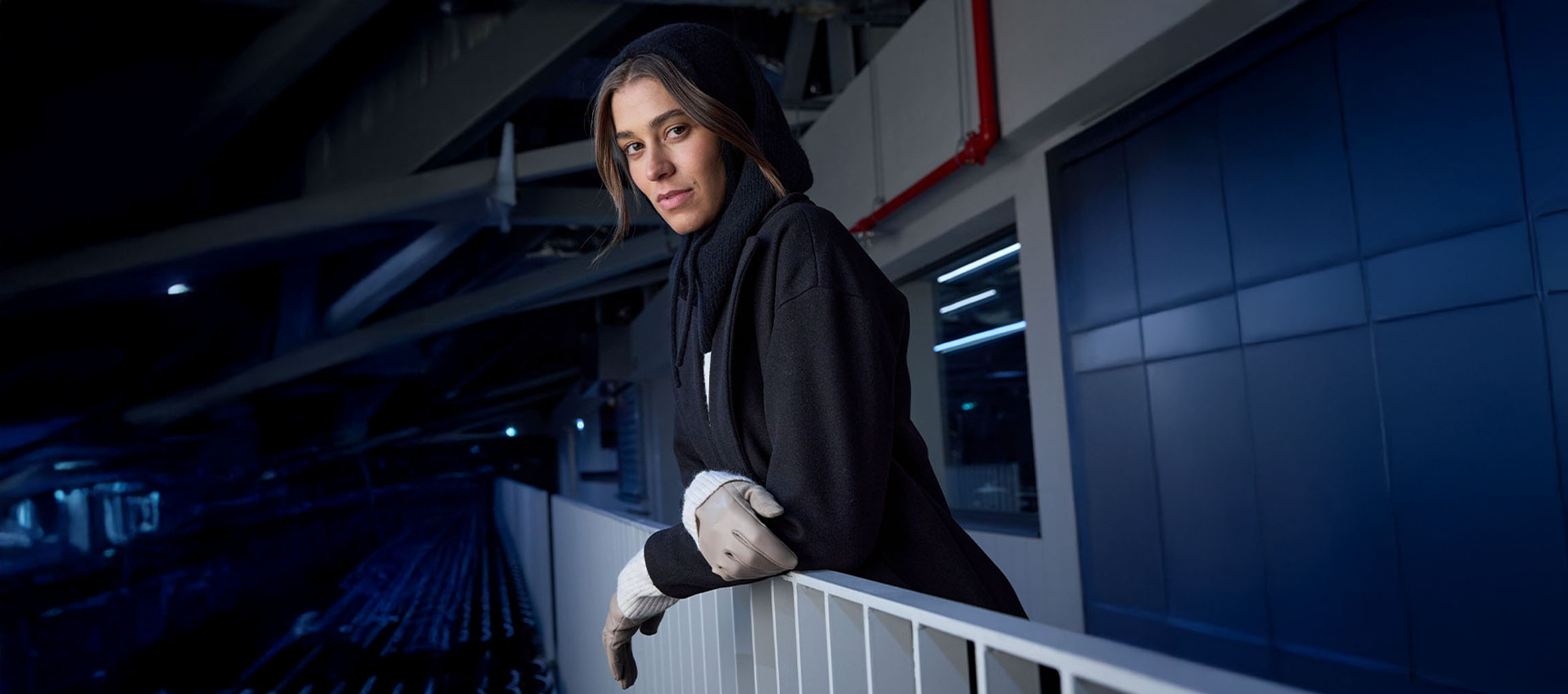 Femeie în palton negru și mănuși, sprijinită de o balustradă într-un cadru urban.