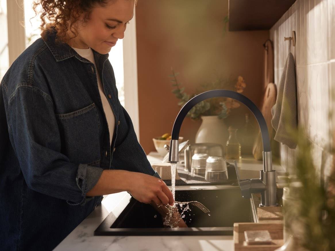 O femeie spală tacâmuri la o chiuvetă modernă cu baterie neagră.