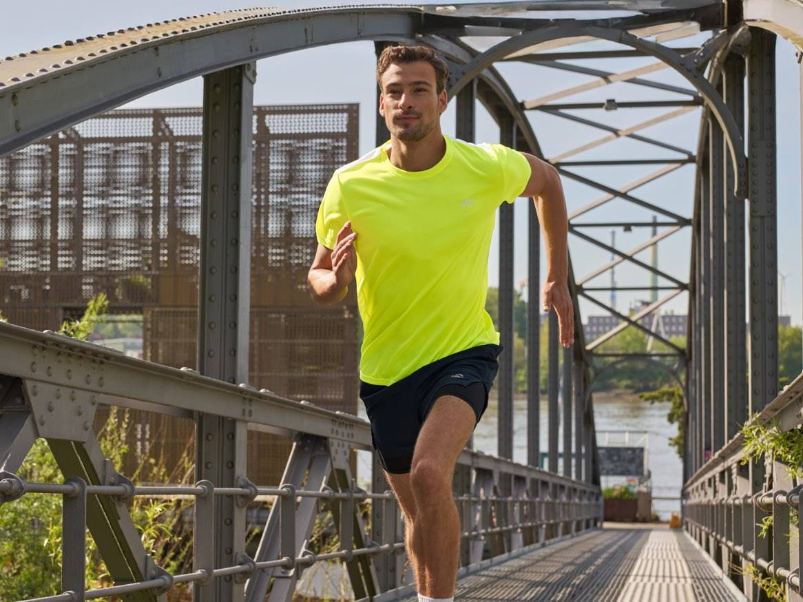 Bărbat în tricou neon și pantaloni scurți negri alergând pe un pod.