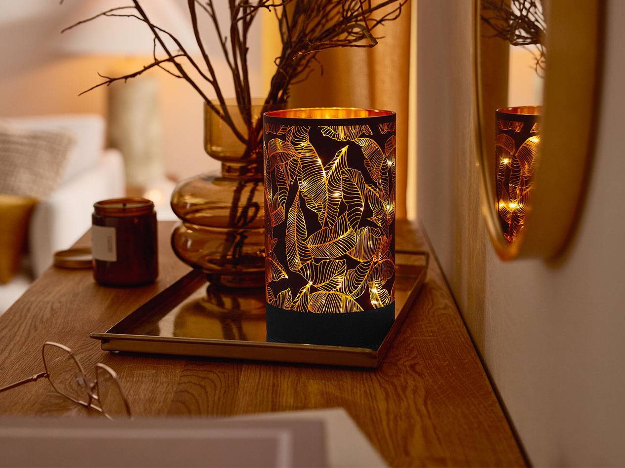 O lanternă LED decorativă cu model de frunze, o vază cu crengi și o lumânare pe o masă de lemn.
