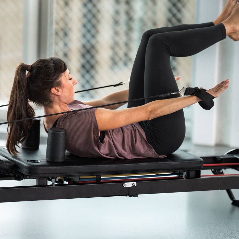 Femeie care face Pilates pe un aparat reformer, purtând echipament sportiv negru.