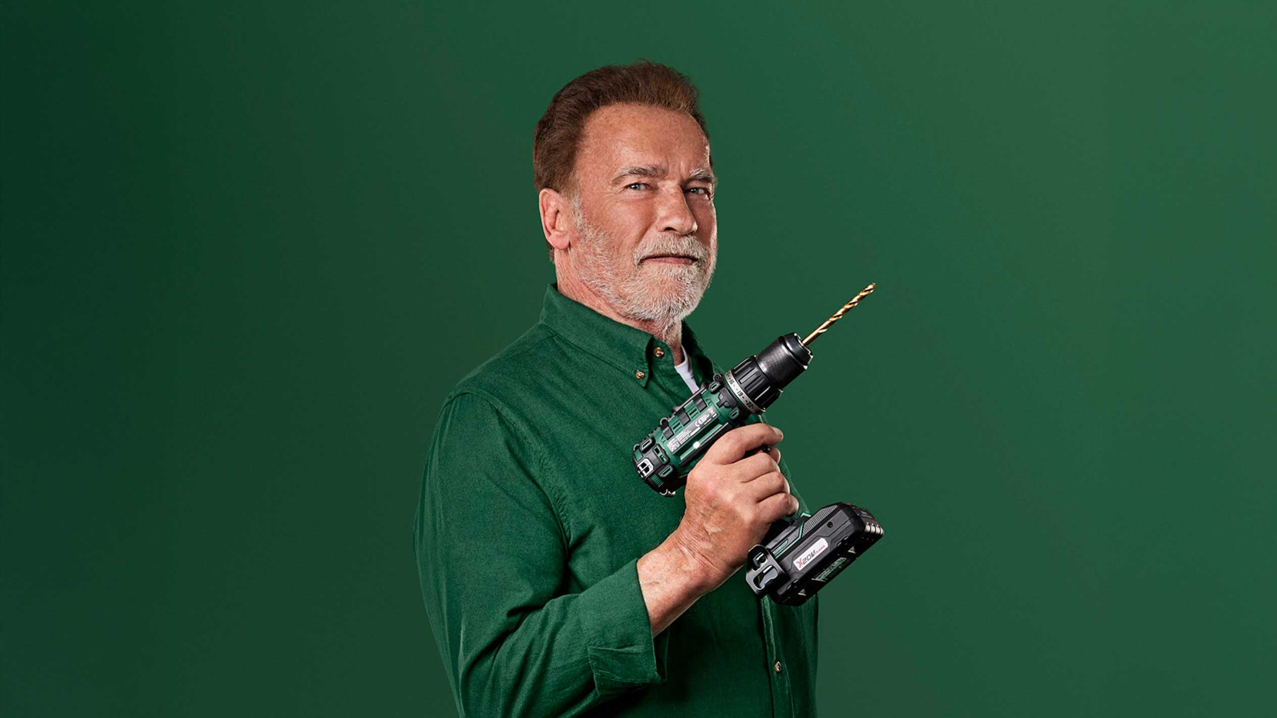 Arnold Schwarzenegger ține o bormașină verde fără fir cu un burghiu auriu.