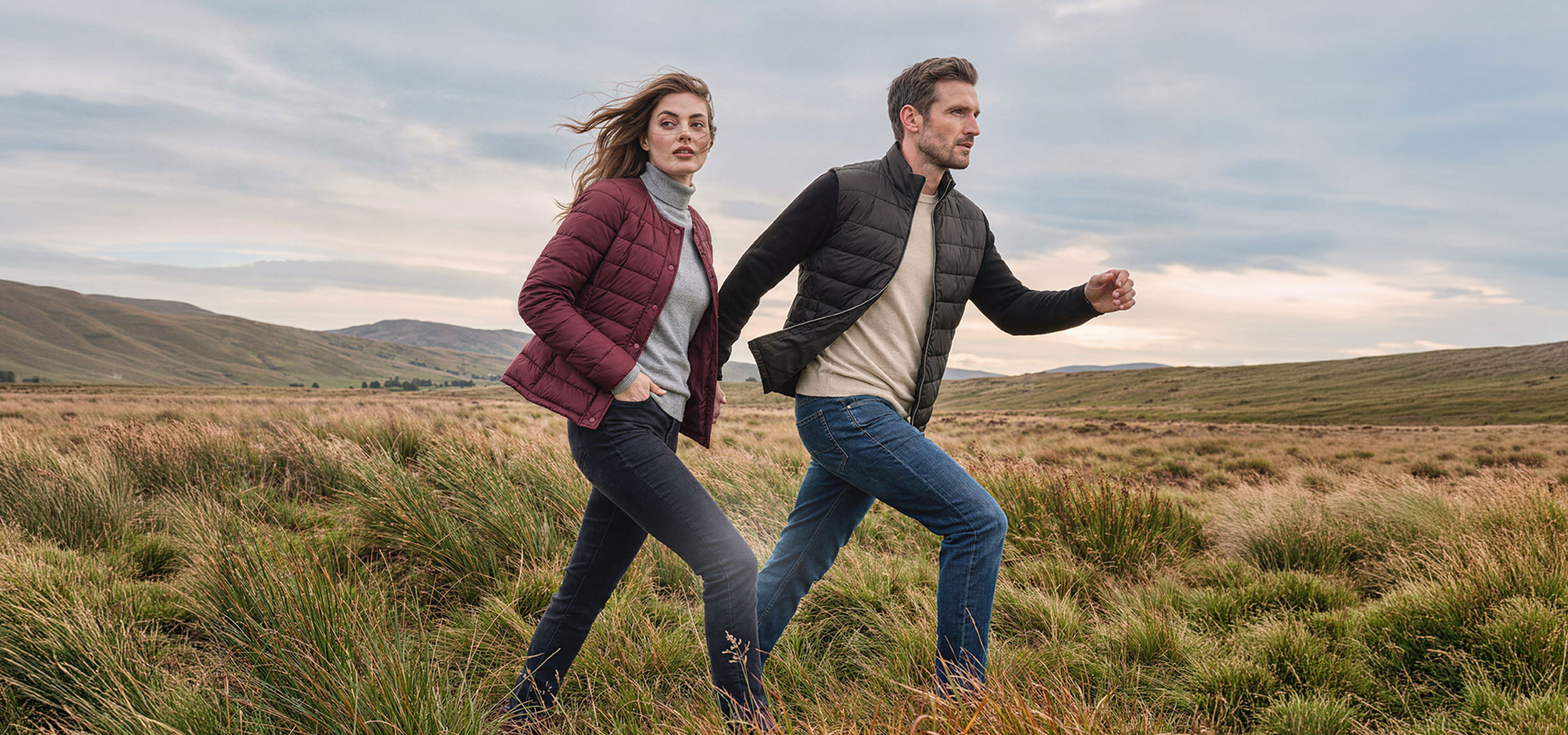 Generat de AI: Paar in leichten Jacken und Jeans geht durch eine grasbewachsene Hügellandschaft.