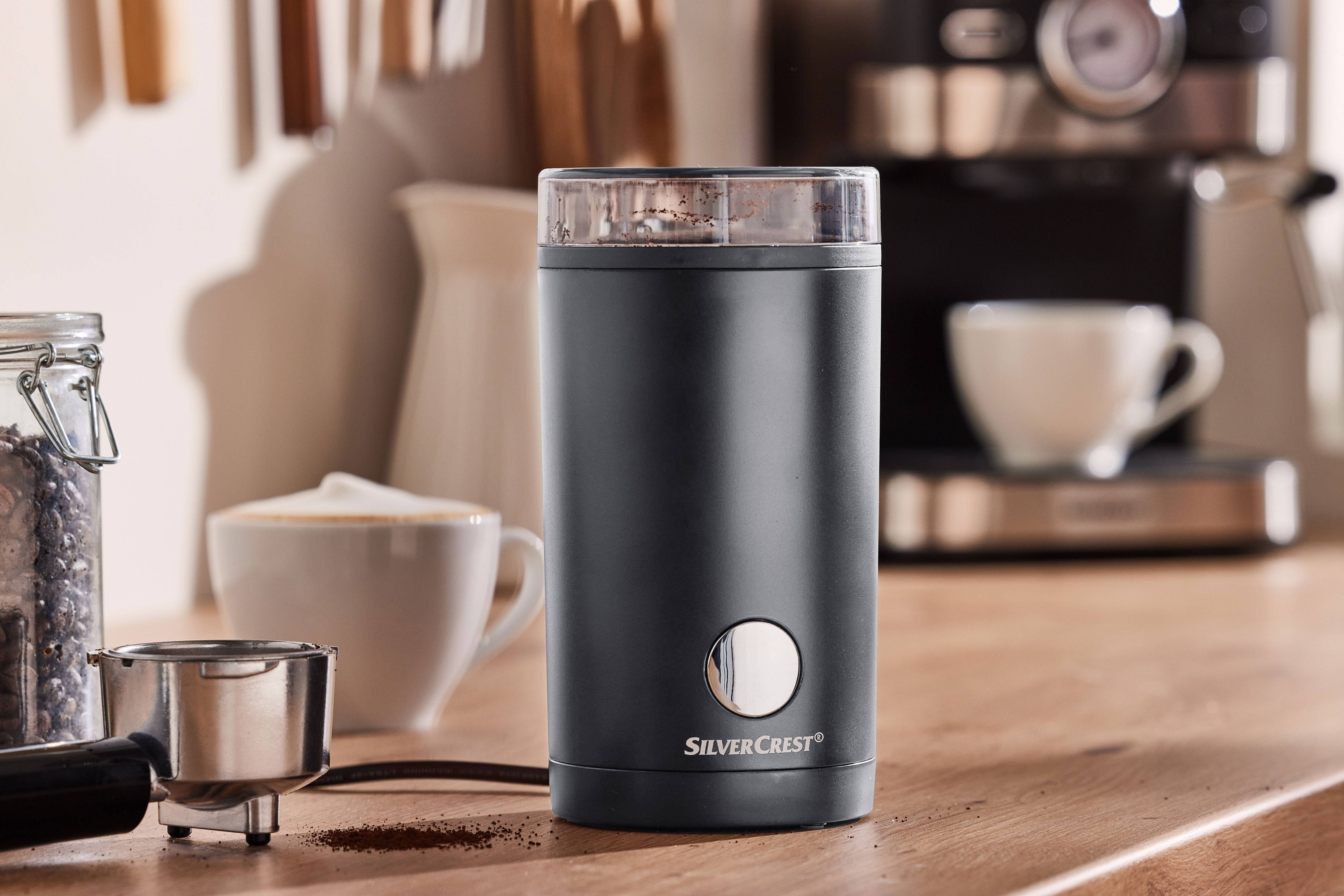 Râșniță neagră de cafea cu cafea măcinată, aparat de cafea și cești pe o masă de lemn.