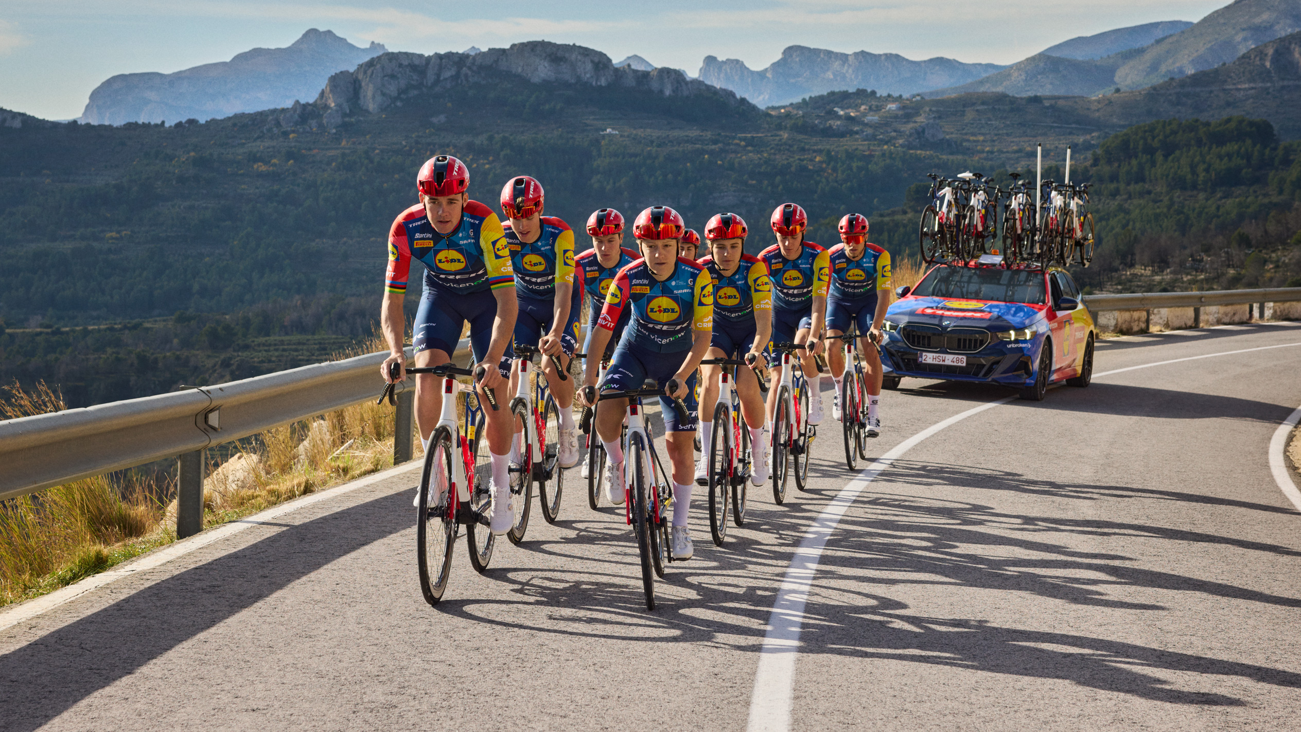 Cicliști în echipamente cu logo-ul unui magazin alimentar, pedalând pe un drum de munte, urmați de o mașină.