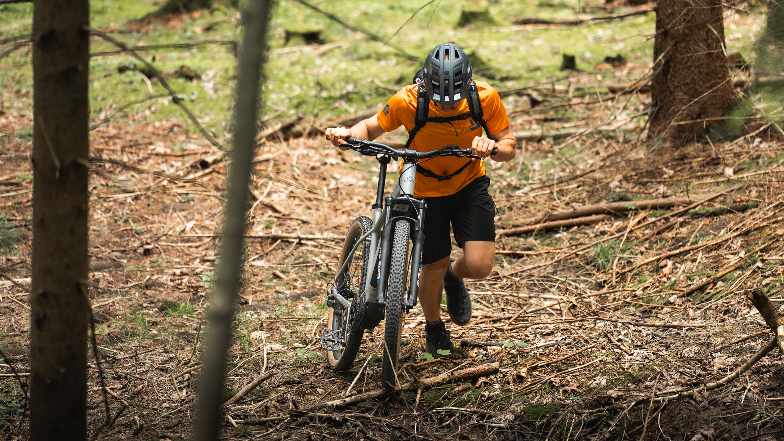 Un ciclist cu tricou portocaliu împinge o bicicletă electrică de munte printr-o pădure.