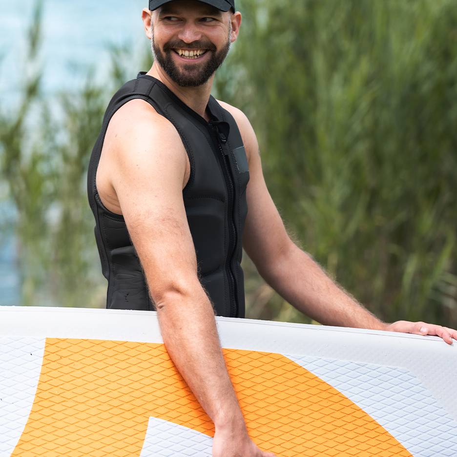 Bărbat zâmbitor în vestă de salvare neagră, ținând o placă de paddleboard.