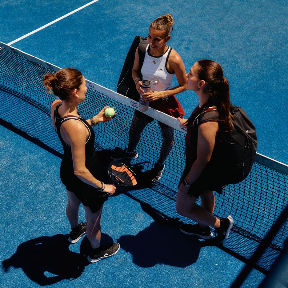 Agostina Bigotti și două colege de joc, în echipament sportiv CRIVIT, stau împreună la fileul de padel pe un teren albastru, după meci.