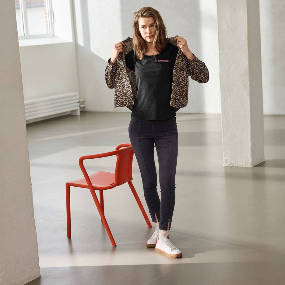 Femeie în tricou negru cu text, blugi și jachetă cu imprimeu leopard, stând lângă un scaun roșu.