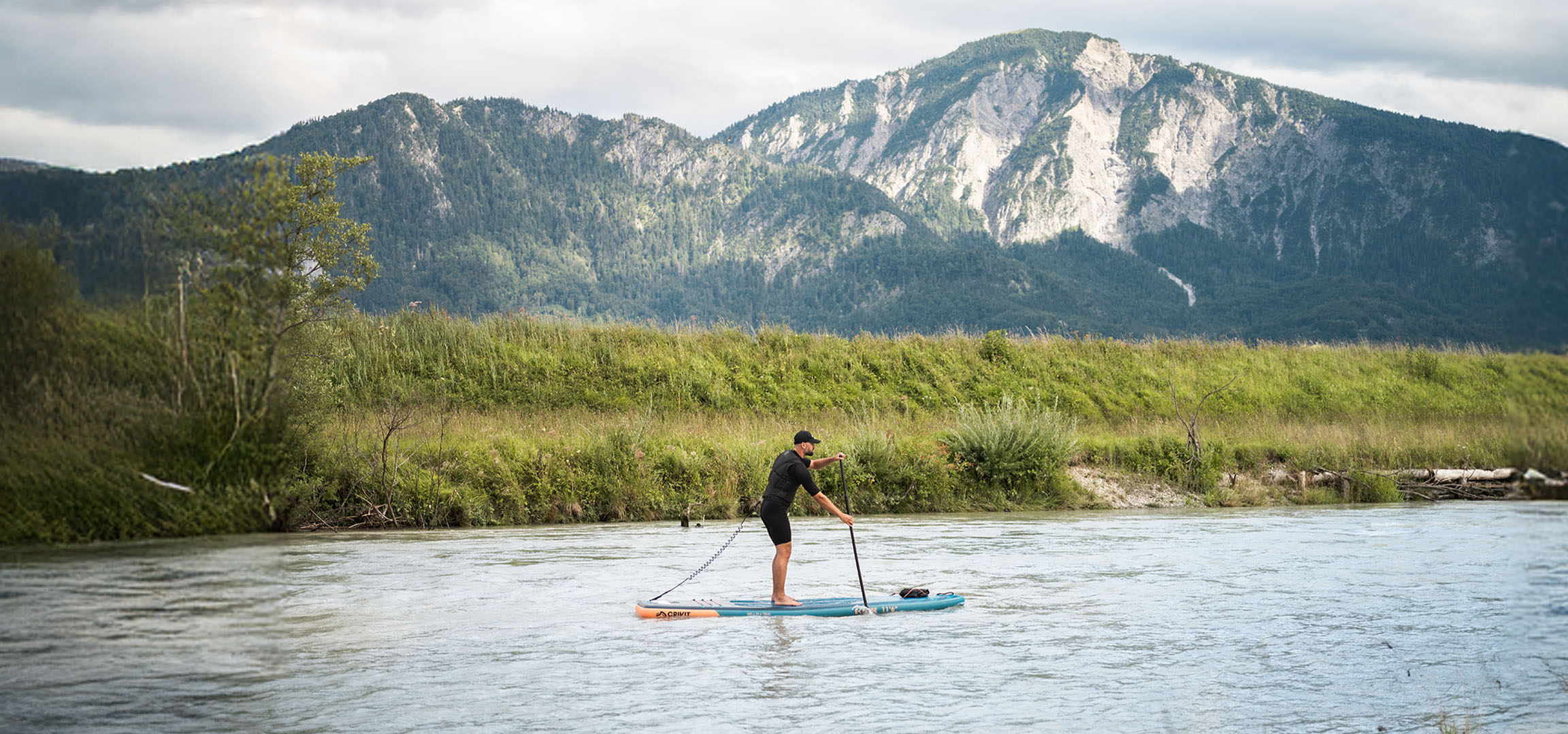 Bărbat pe paddleboard pe un râu cu munți și vegetație luxuriantă în fundal.