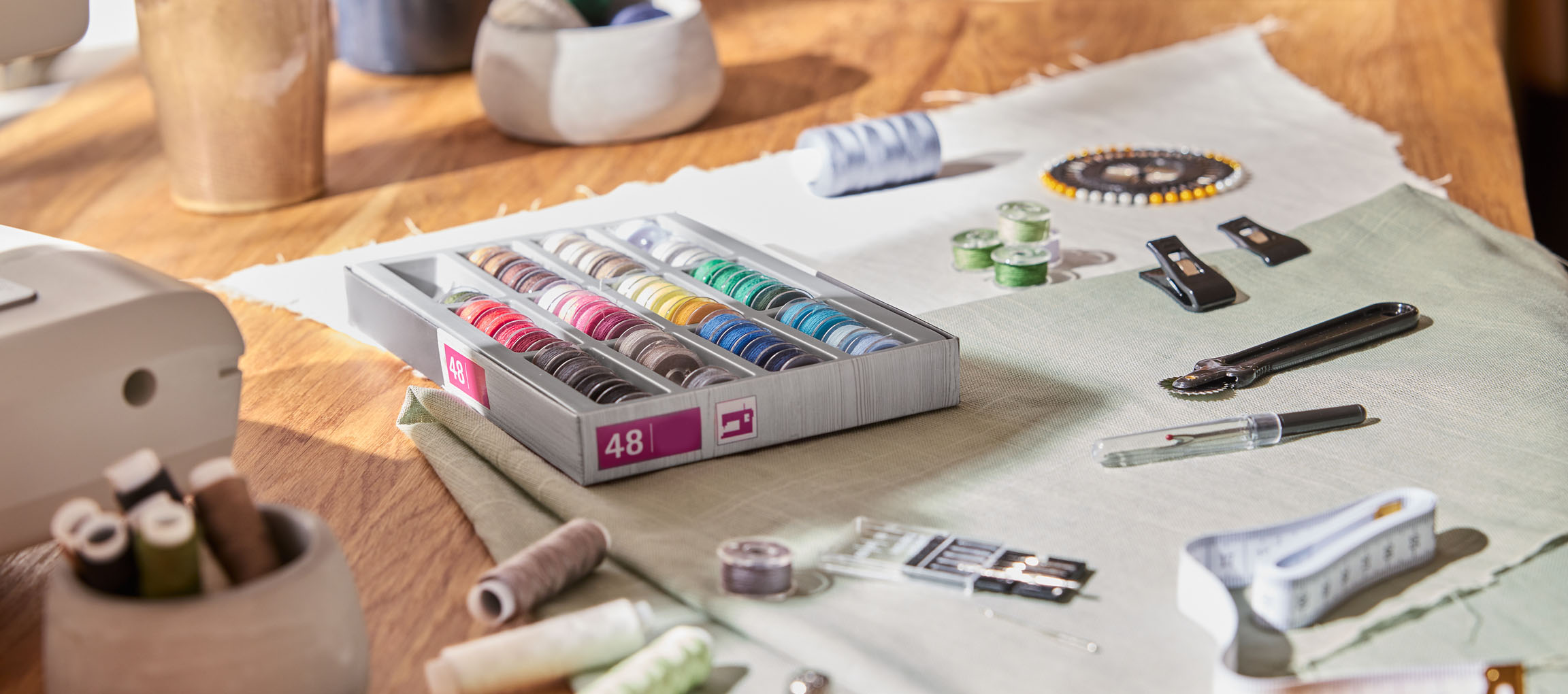 Accesorii de cusut pe o masă de lemn, inclusiv un organizator de ațe cu 48 de bobine, material și diverse unelte.