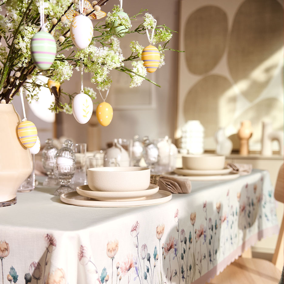 Ostertisch gedeckt mit floraler Tischdecke, Keramikgeschirr und Ästen mit Ostereiern.
